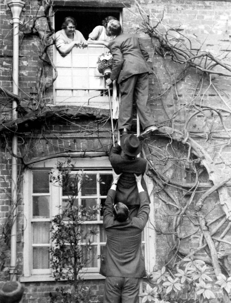 A black and white photo from the 1930s of two women at the first floor window of a brick house with vines. A man is standing on a ladder and kissing one of the women, while the ladder is held by two other men. 